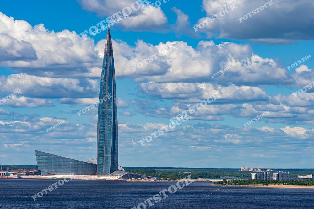 Современный высокий небоскреб в виде остроконечного стеклянного здания доминирует в ландшафте, на фоне облаков и синего неба. Здание стоит на берегу широкой водной поверхности, чьи волны отражают его силуэт. На заднем плане видны леса и меньшие постройки, придающие сцене перспективу и контраст современной архитектуры. Фотография демонстрирует небоскреб, возвышающийся на берегу Финского залива в Санкт-Петербурге. Современное высотное здание отражает деловой ритм города и развитие бизнеса в регионе. На фоне яркого голубого неба архитектура небоскреба выглядит особенно впечатляюще, подчеркивая гармонию современной урбанистики и природного окружения. Солнечная погода придает кадру дополнительную яркость и привлекательность.